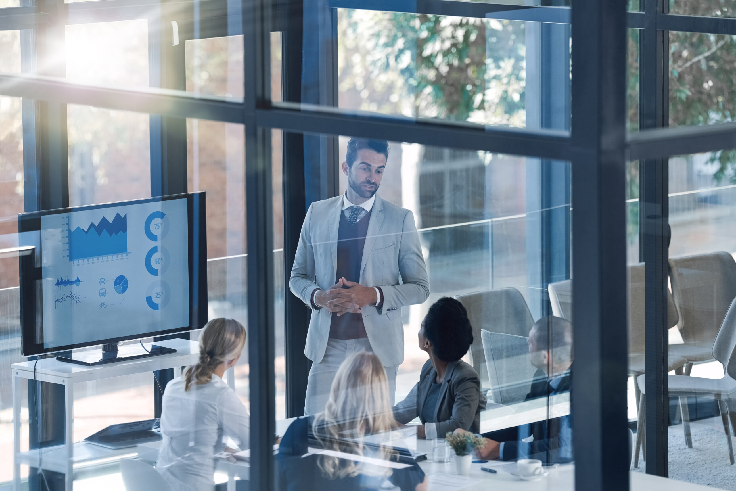 Meeting, presentation and business people with manager on screen statistics, data analytics and charts by window. Speaker or man speaking to employees with infographics, revenue and profit on monitor.